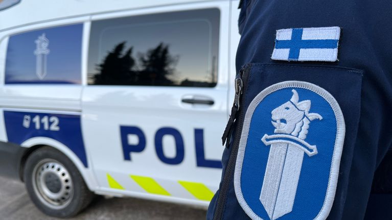 Oikealla etualalla poliisin hihamerkki ja Suomen lippu hihassa. Taustalla poliisiauto. Tiivis rajaus.