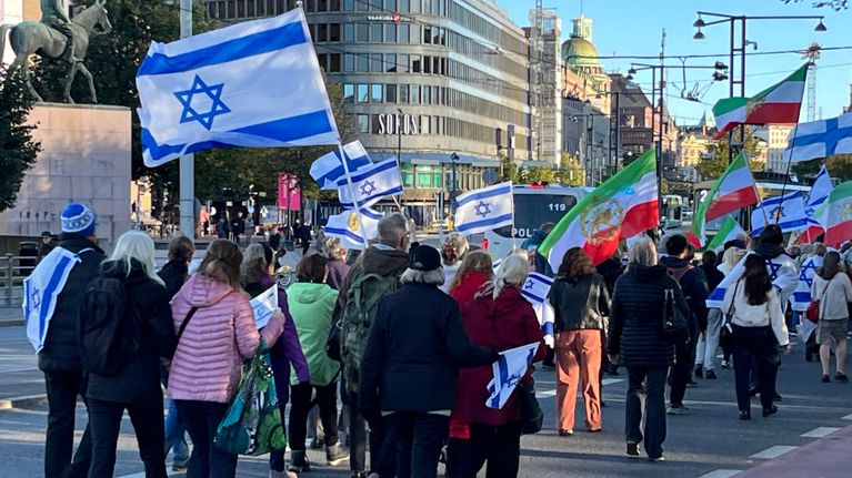 Ihmisiä kulkee kadulla kantaen Israelin ja Iranin lippuja kaupungin keskustassa, taustalla patsas ja liikennettä.