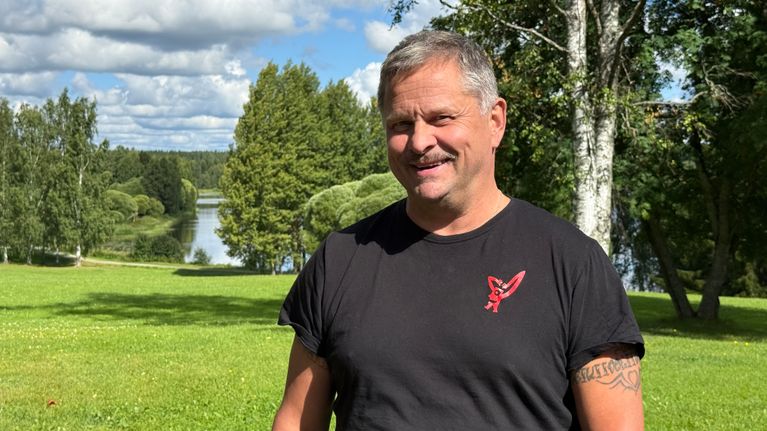 Näyttelijä Ismo Apell virnistää kameraan katsoen. Taustalla on vehreää nurmikkoa ja tyyni vesistö.