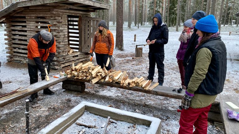 Five people stand around in a semi-circle watching a man demonstrating woodcarving in a forest with a woodshed behind.