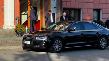 Svart audi står parkerad utanför hotellentré, säkerhetsvakter rör sig runt bilen.