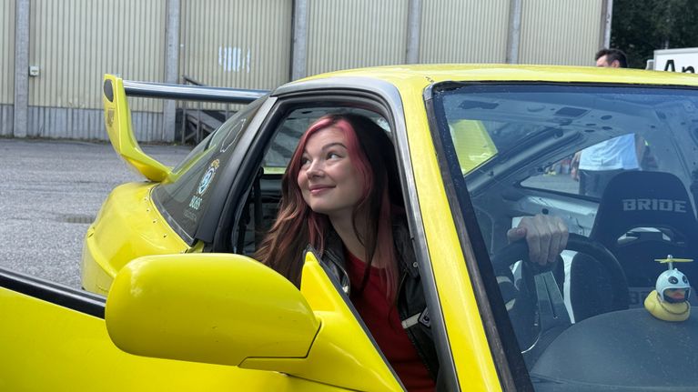 Näyttelijä Emma Kilpimaa kurkkaa hymyillen keltaisesta autosta kuljettajan paikalta. Auton kojelaudalla on keltainen kylpyankka.