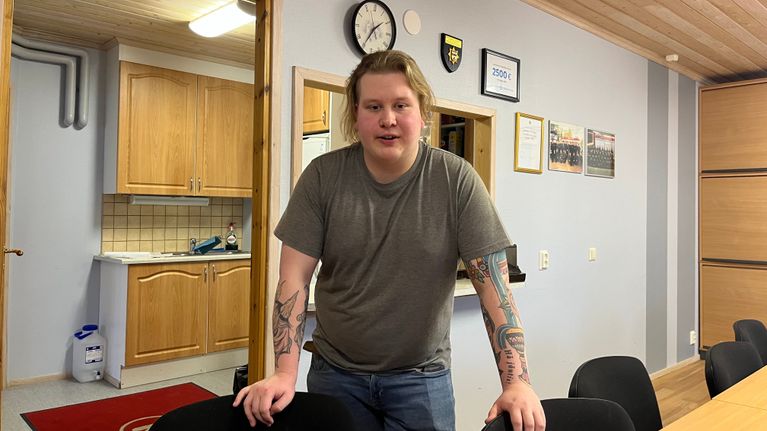 Mies seisoo toimistotilassa nojaten pöytään, hänen ympärillään on tuoleja.