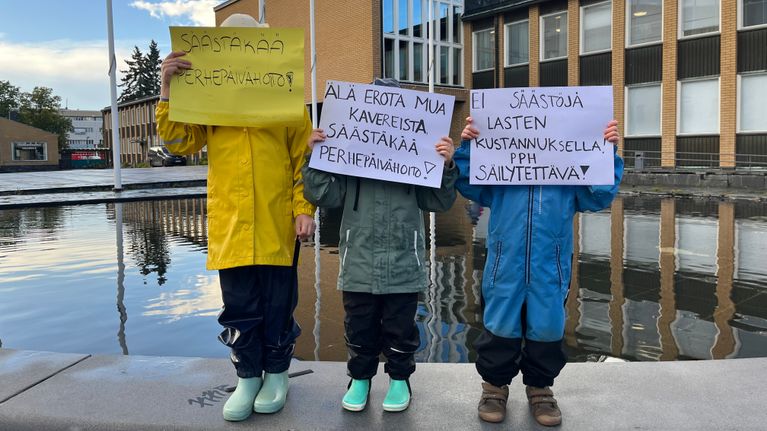 Kolme lasta seisoo vesialtaan laidalla ja pitää kasvojensa edessä papereita, joissa puolustetaan perhepäivähoitoleikkauksia.