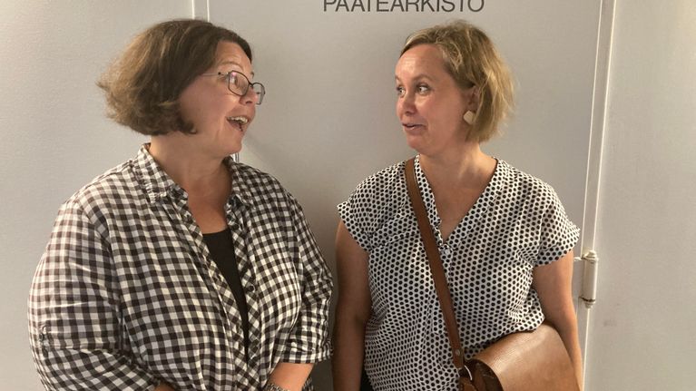 Kaksi naista katsoo toisiinsa. He seisovat oven edessä. Siinä tulee päätearkisto.