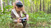 Woman kneeling in the forest wearing a goggle headset and holding a spectral imaging camera by a mushroom on the forest floor.