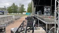 Esiintymislava, jonka päällä ja ympärillä on kuljetuslaatikoita. Lavan edessä on aitarakennelmia. Taka-alalla näkyy puita ja järveä. 