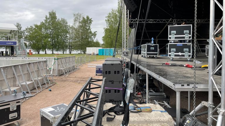 Esiintymislava, jonka päällä ja ympärillä on kuljetuslaatikoita. Lavan edessä on aitarakennelmia. Taka-alalla näkyy puita ja järveä. 