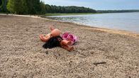 En docka ligger övergiven på en sandstrand vid en sjö, omgiven av natur.