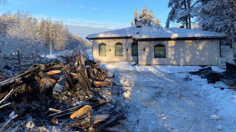 Kuvassa on valkoista tiiltä oleva rakennus ja edessä on röykkiä puuta ja romua tulipalon jäljiltä