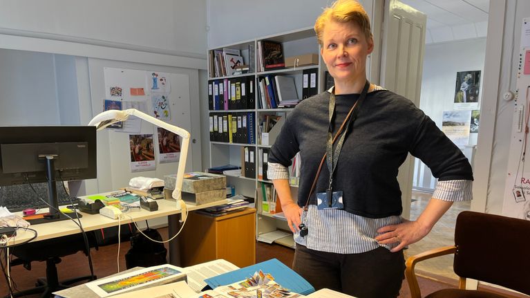Nainen seisoo pöydän vieressä kädet vyötäröllä. Pöydällä on mappeja ja kirjoja.