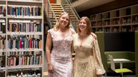 Två personer i ljusa klänningar står bredvid varandra i ett modernt bibliotek med bokhyllor och sittplatser.