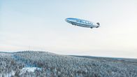 A silver airship with the word KELLUU on the side above a snowy, forested landscape.