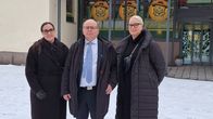 Kolme henkilöä seisovat lumisessa maisemassa talvivaatteet yllään, taustalla kauppakeskus Chydenia, jonka ikkunoissa värikkäitä kuvia.
