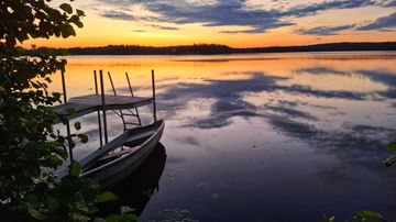 Järvimaisema, jossa laituri ja vene, järvi heijastaa iltaruskon värejä, taustalla metsäinen ranta.