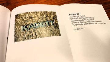 t foto av ett Kadett-emblem på en stenig yta, bredvid texten "Näyte 26" och beskrivning på finska.