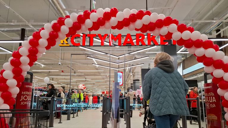 Nainen kävelee Citymarketin porteista. Nainen työntää lastenrattaita. Porttien yläpuolella on ilmapalloista tehty kaari.