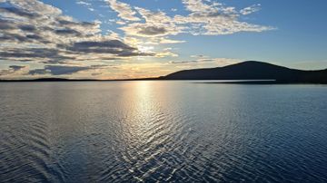 Aurinko on laskemassa järven yllä, taustalla näkyy matalia pilviä ja loiva mäki horisontissa.