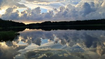 Pilvinen taivas ja metsän reunustama järvi, jonka tyyni pinta heijastaa pilvimuodostelmia.