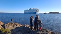 Massive white cruise ship on the sea, people in the foreground on rocky coast looking at the vessel sail past. Islands and two other boats are seen by the ship.