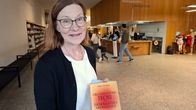 En person med glasögon, vit skjorta och mörk tröja håller upp en bok med titeln "Organisationsteori och teknokratiskt medvetande" av Mats Alvesson. Bilden är tagen i ett bibliotek.