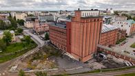 Stort tegelhus med flera våningar intill ett område med rivningsarbete, omgiven av bostadshus och järnvägsspår.