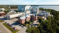 Sjukhusbyggnader med flera våningar och parkering, omgivna av träd och vatten i bakgrunden.