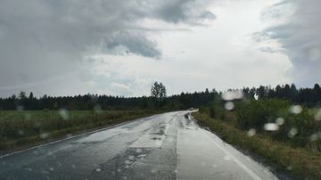 Märkä maantie kaareutuu metsän reunustamana harmaan pilvistä taivasta vasten, vesipisaroita näkyy tien pinnalla.