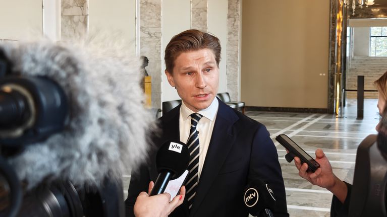 Man in dark suit jacket, white shirt and striped necktie speaking to reporters holding microphones.
