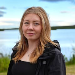 Nuori nainen seisoo ulkona järven rannalla, taustalla näkyy vesistöä, rantakasvillisuutta ja pilvinen taivas.
