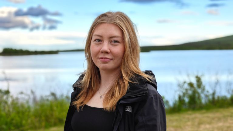 Nuori nainen seisoo ulkona järven rannalla, taustalla näkyy vesistöä, rantakasvillisuutta ja pilvinen taivas.