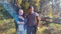 Två personer står utomhus framför bikupor, ena personen håller en katt och de omges av höstträd.