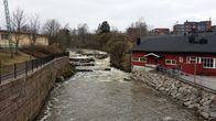 En å rinner genom ett stadsmiljöområde med en röd byggnad till höger och stenmur längs vänster sida.
