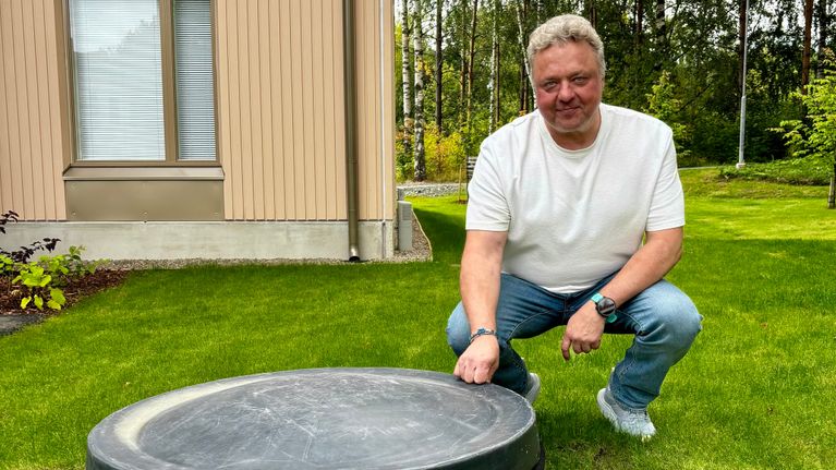 Mies on kyykyssä nurmikolla olevan mustan säiliönkannen äärellä. 