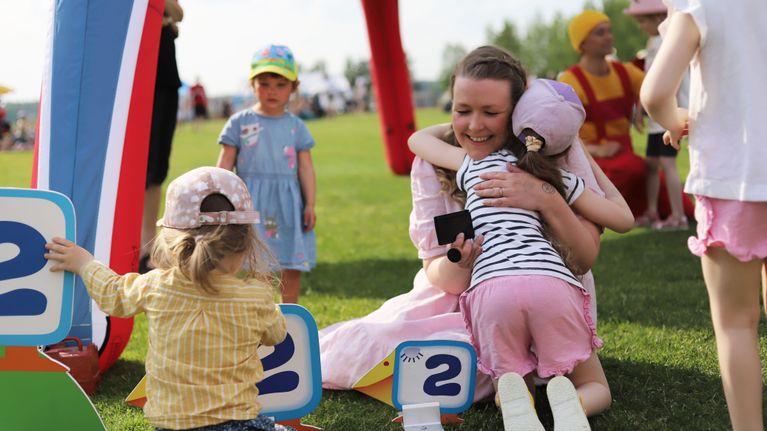 Pikku Kakkosen juontaja Susanna istuu nurmikolla ja halaa iloisena lippispäistä lasta. Vieressä on lisää pieniä lapsia sekä Pikku Kakkosen logosta tuttuja pahvisia lintuhahmoja.