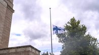 Suomen lippu puolessa tangossa kivirakennuksen edustalla, taustalla puu ja pilvinen taivas.