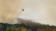 Helikopteri pudottaa vettä metsäpaloon savun noustessa metsän yläpuolella, etualalla talojen kattoja.