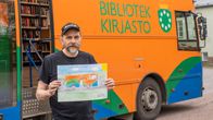 En person håller upp en teckning framför en orange bokbuss med texten "Bibliotek Kirjasto" utanför en byggnad.