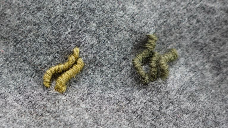 Kuvassa on yksityiskohta harmaasta villakankaasta, jossa on kaksi pientä ryppyä tai muodostelmaa vihreää lankaa.