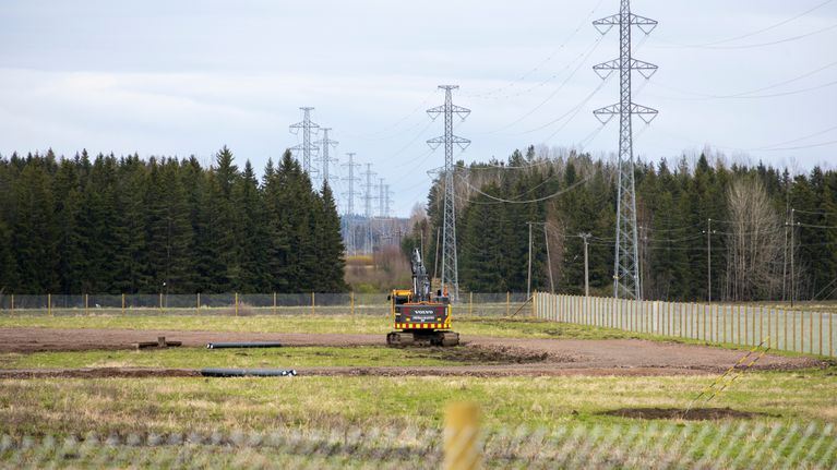 An excavator in the middle of a field, with trees and high voltage power lines seen in the distance.