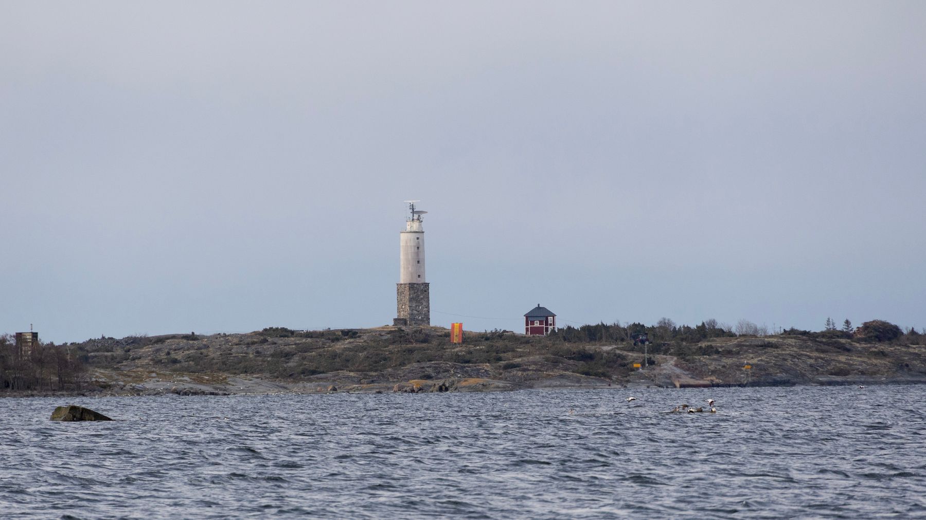 Majakka, punainen rakennus ja linjataulu lähes puuttomalla saarella. Etualalla merta.