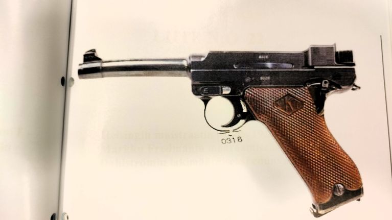 En pistol med brunt handtag och metallisk pistolram ligger på ett vitt underlag. Siffran "0318" syns under avtryckaren.
