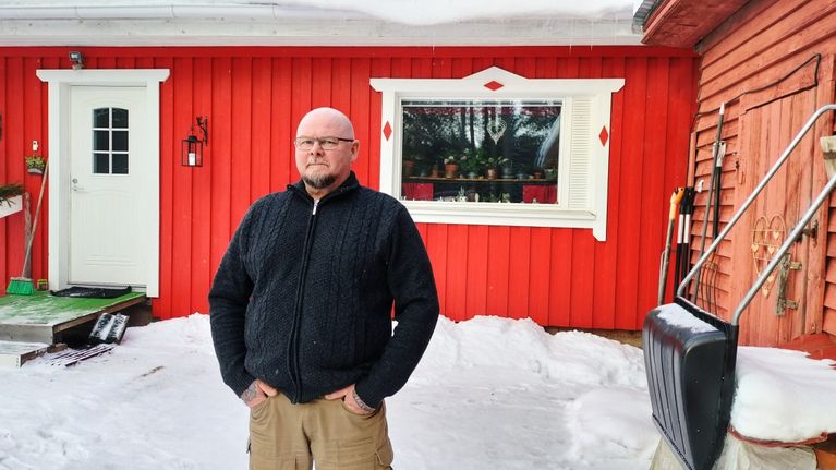 Peter Dahlström står framför ett rött hus med vita detaljer, snö på marken och redskap lutade mot väggen.