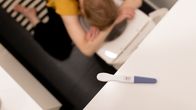 Nainen oksentaa pöntöllö, etualalla positiivinen raskaustesti.