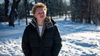 En ung person står ute i ett vintrigt landskap med snö omkring sig. Personen bär en mörk vinterjacka och det är soligt väder. 