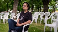 Mustaan t-paitaan ja farkkuihin pukeutunut mies istuu puistossa valkoisella muovipenkillä ja katsoo yläilmoihin. Miehellä tummasankaiset silmälasit ja hattu, taustalla vehreää puistoa.