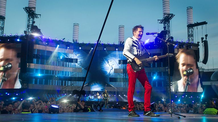 Rockyhtye Muse lavalla. Kitaristi, isot videoscreenit taustalla.