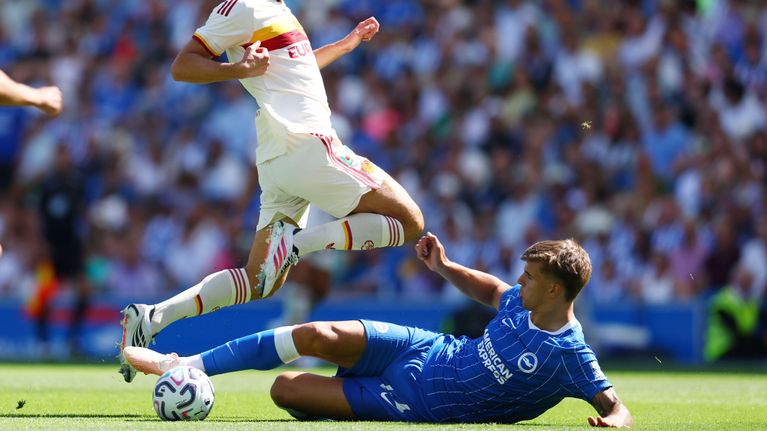 En spelare i vitt blir tacklad av en spelare i blått på en fotbollsplan, inför publik.