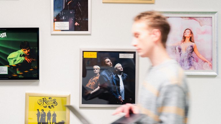 Ylen toimittaja kävelee seinän editse, jossa näkyy UMK-artistien kuvia.