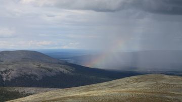 Pilvinen maisema tunturin huipulta, taustalla näkyy sateenkaari ja metsäisiä laaksoja.